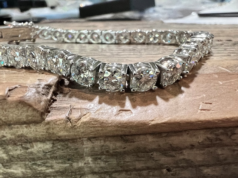 K&ouml;nnte beinhalten: Ein funkelndes Tennisarmband mit zahlreichen rund geschliffenen Diamanten, die in einem silberfarbenen Metall gefasst sind. Die Diamanten sind eng aneinander gesetzt und erzeugen eine durchgehende Linie. Das Armband liegt auf einer Holzoberfl&auml;che.