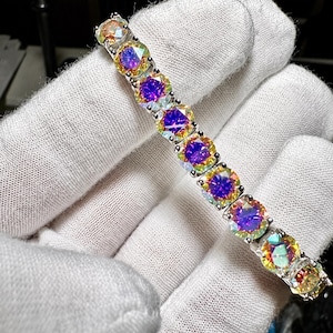 May include: A tennis bracelet featuring a series of round, faceted iridescent stones set in a silver-toned metal frame. Each stone displays a vibrant color shift, transitioning from deep purple centers to bright yellow and green edges in a repeating pattern.