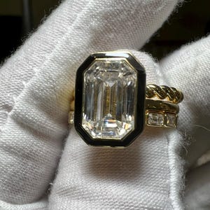 May include: A gold ring with a large, emerald-cut diamond set in a black enamel bezel. Additional gold rings with diamonds and a twisted rope design are stacked alongside the main ring. The rings are displayed on a white surface.