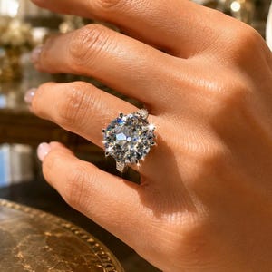 May include: A silver-toned ring featuring a large, round, brilliant-cut clear gemstone set in a multi-prong setting. The band is slender and metallic, showcasing a reflective surface that catches the light, creating a bright, sparkling appearance.