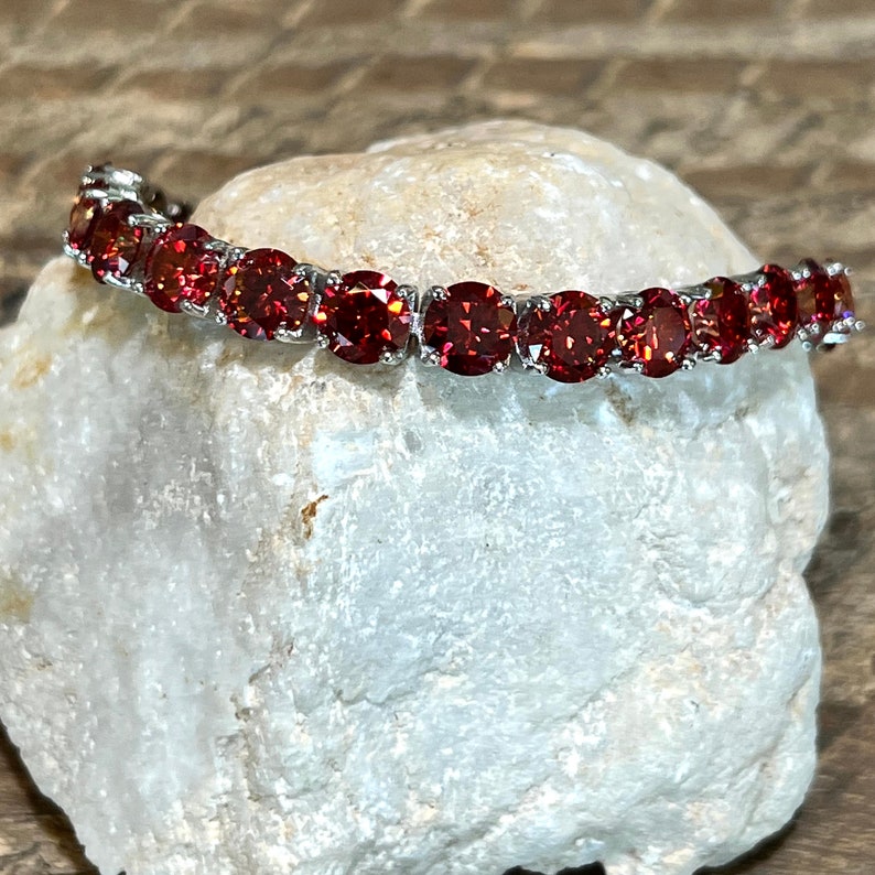 May include: A silver bracelet with a row of red gemstones. The bracelet is on a white rock.