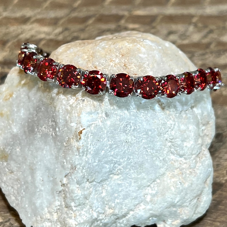 May include: A silver bracelet with a row of red gemstones. The bracelet is on a white rock.