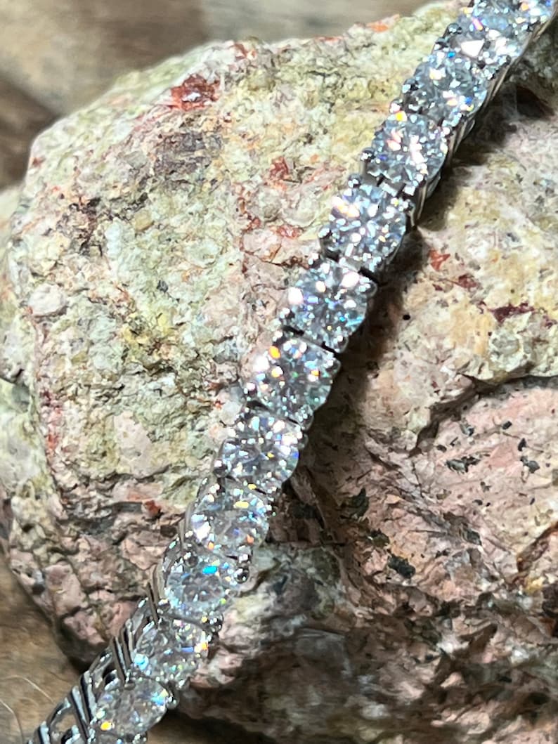 K&ouml;nnte beinhalten: Ein silbernes Tennisarmband mit zahlreichen runden, klaren Edelsteinen. Das Armband wird auf einer strukturierten, hellbraunen und grauen Felsfl&auml;che pr&auml;sentiert. Die Edelsteine funkeln und reflektieren das Licht.