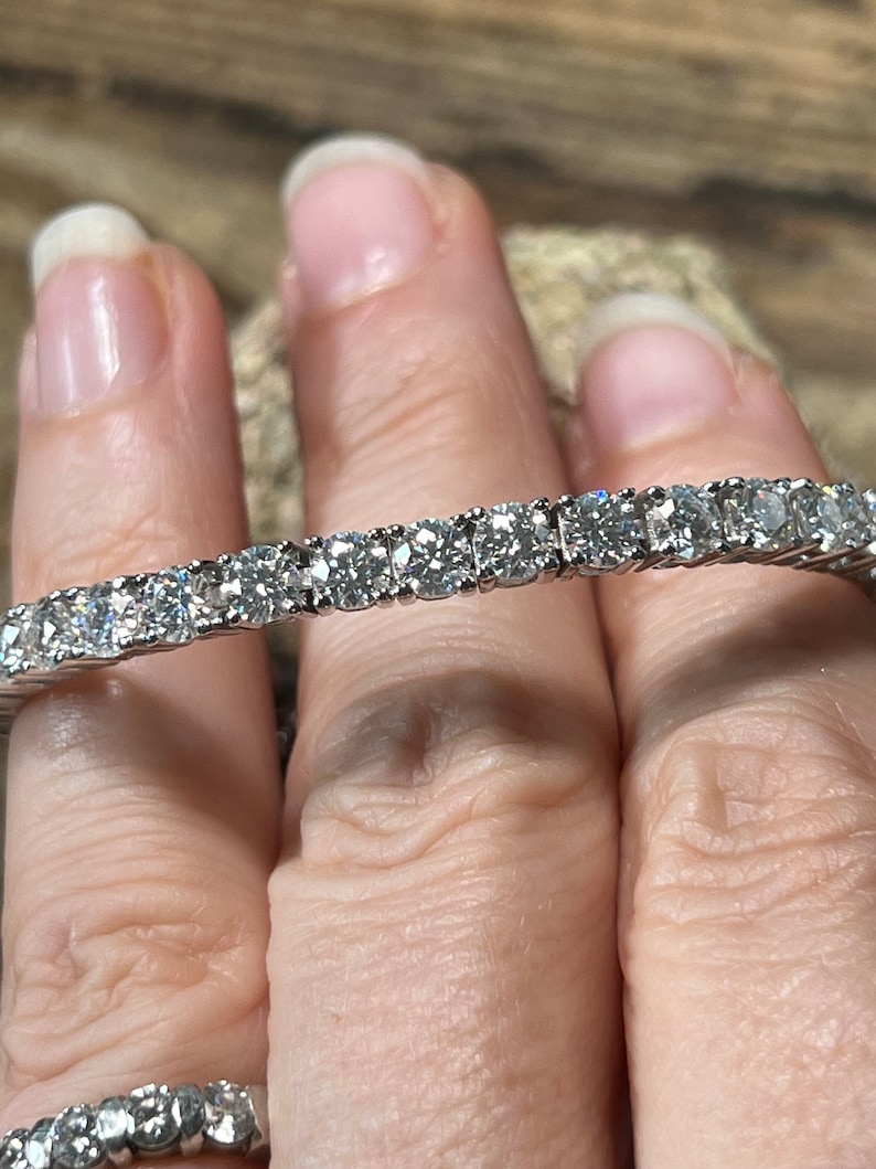 K&ouml;nnte beinhalten: Nahaufnahme eines Tennisarmbands mit einer Reihe funkelnder, rund geschliffener Diamanten, die in einem silbernen Band gefasst sind. Das Armband wird von einer Person gehalten. Die Diamanten reflektieren das Licht.
