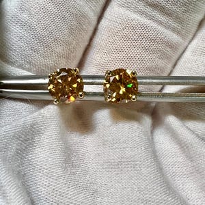 May include: A pair of round, golden-brown gemstone stud earrings. The earrings are set in gold and are displayed on a silver metal tool against a white fabric background. The gemstones have a brilliant cut.