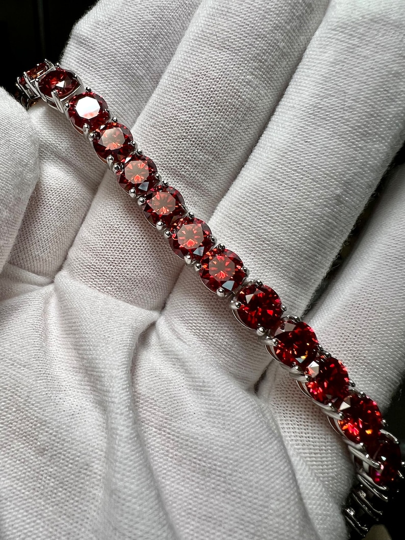 May include: A silver tennis bracelet with a row of red round gemstones.