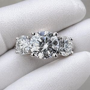 Op de afbeelding: Een ring met drie stenen met een grote, ronde diamant in het midden en twee kleinere diamanten aan weerszijden. De ring is gezet in een wit metaal, waarschijnlijk platina of witgoud, en wordt getoond tegen een witte achtergrond.