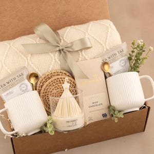 May include: A gift box containing a cream-colored cable knit blanket tied with a sage green ribbon, two white ribbed ceramic mugs, two gold-toned spoons, two tea packets labeled Tea With Tae, a milk chocolate bar, woven coasters, and a candle.