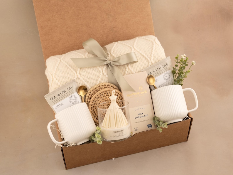 May include: A gift box containing a cream-colored cable knit blanket tied with a sage green ribbon, two white ribbed ceramic mugs, two gold-toned spoons, two tea packets labeled Tea With Tae, a milk chocolate bar, woven coasters, and a candle.