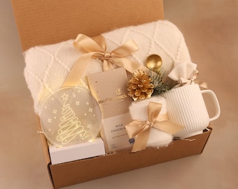 May include: A gift box filled with a white blanket, a mug, a chocolate bar, a decorative Christmas tree light, and other items. The box is tied with gold ribbons and includes a gold spoon and pinecone.