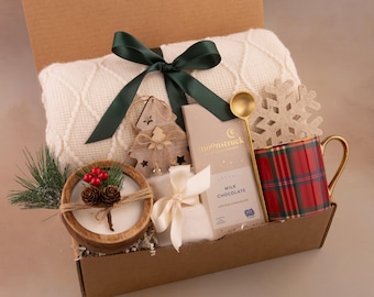 May include: A gift box filled with a cream-colored knitted blanket, a red plaid mug, a wooden candle, a chocolate bar, and a decorative snowflake. The box is tied with a dark green ribbon, creating a cozy, festive theme.