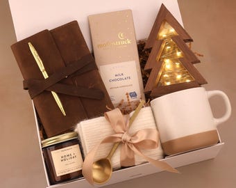 Puede incluir: Una caja de regalo llena de un diario de cuero marrón y un bolígrafo dorado, una tableta de chocolate con leche, un árbol de Navidad de madera con luces de hadas, una taza blanca y beige, una vela, una cuchara dorada y una manta color crema.