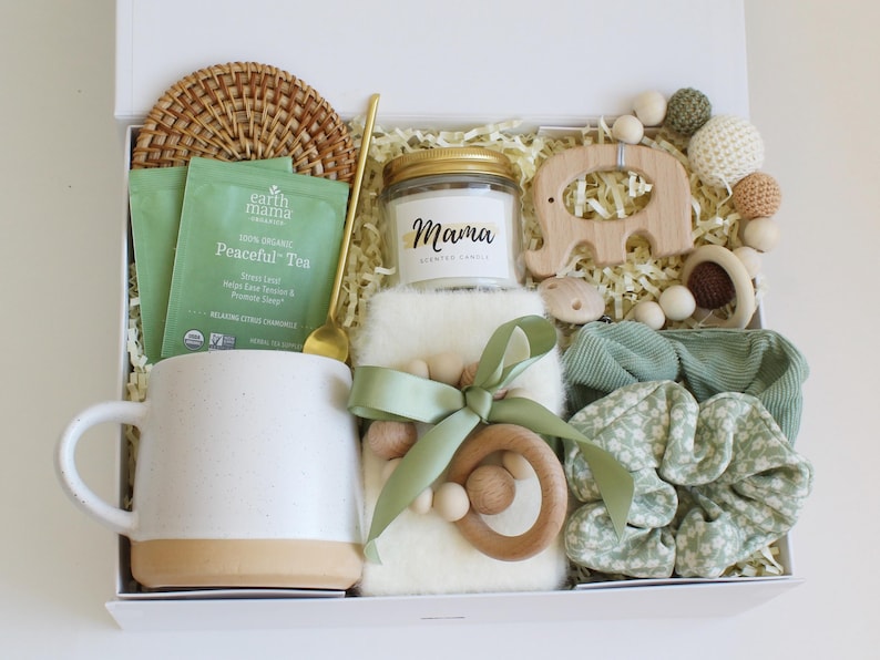Puede incluir: Una caja de regalo blanca llena de varios art&iacute;culos. Incluye una taza, t&eacute;, una vela con el texto "Mama", un elefante de madera, un mordedor de madera, coleteros y una cuchara dorada. Los art&iacute;culos son en tonos blanco, beige y verde.