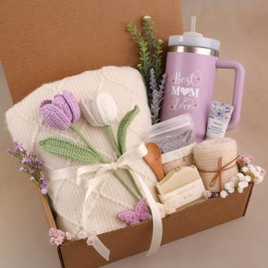 May include: A gift box filled with a white blanket, a purple tumbler with the text "Best Mom Ever", a bar of soap, a wooden spoon, and a bundle of lavender. The box also contains crocheted purple and white tulips.