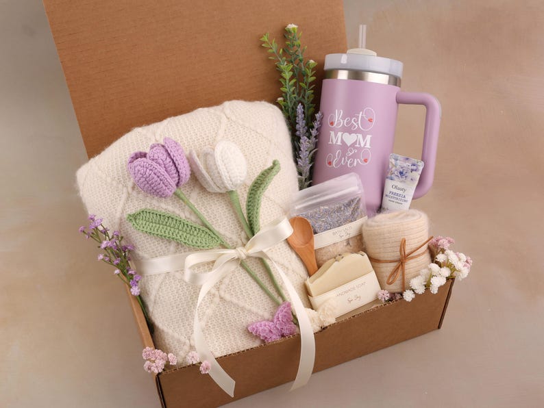 May include: A gift box filled with a white blanket, a purple tumbler with the text "Best Mom Ever", a bar of soap, a wooden spoon, and a bundle of lavender. The box also contains crocheted purple and white tulips.