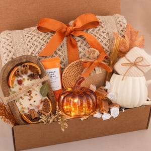 May include: A gift box containing a cream-colored knit blanket with an orange ribbon, a wooden bowl candle with dried orange slices and cinnamon, a tube of vegan hand cream, a glass pumpkin, a white pumpkin-shaped mug, and wrapped candies.