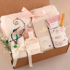May include: A gift box filled with a white blanket, a floral mug with "MOM" text, chocolate, tea, bath salts, and handmade soap. A stained glass hummingbird and pink ribbon add decorative touches.