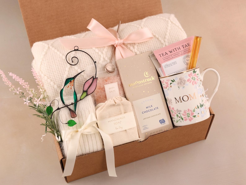 May include: A gift box filled with a white blanket, a floral mug with "MOM" text, chocolate, tea, bath salts, and handmade soap. A stained glass hummingbird and pink ribbon add decorative touches.