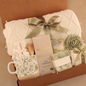 May include: A gift box filled with a cream-colored blanket, a floral-patterned mug, milk chocolate, soap, bath salts, a wooden spoon, and a decorative flower. The box is tied with a sage green ribbon.