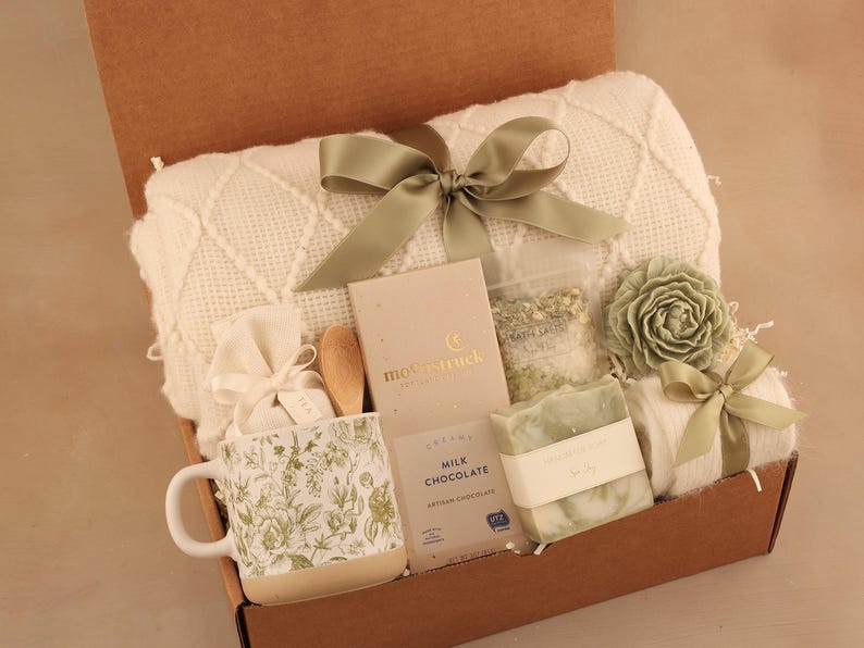 May include: A gift box filled with a cream-colored blanket, a floral-patterned mug, milk chocolate, soap, bath salts, a wooden spoon, and a decorative flower. The box is tied with a sage green ribbon.