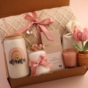 Puede incluir: Una caja de regalo llena de varios art&iacute;culos, incluyendo un vaso de vidrio con un dise&ntilde;o de arco&iacute;ris y la palabra "mama", una barra de chocolate con leche, una vela, un tulip&aacute;n de ganchillo y una manta. Los art&iacute;culos est&aacute;n atados con cintas rosas.