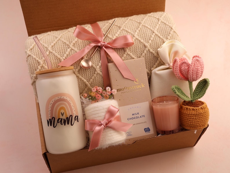 Puede incluir: Una caja de regalo llena de varios art&iacute;culos, incluyendo un vaso de vidrio con un dise&ntilde;o de arco&iacute;ris y la palabra "mama", una barra de chocolate con leche, una vela, un tulip&aacute;n de ganchillo y una manta. Los art&iacute;culos est&aacute;n atados con cintas rosas.