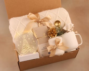 May include: A gift box filled with a white blanket, a mug, a chocolate bar, and a decorative Christmas tree light. The box also contains a gold spoon, a pine cone, and a pair of socks tied with a gold ribbon.