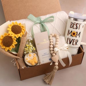 May include: A gift box filled with a white blanket, a tumbler with "BEST MOM EVER" text, a bouquet of crochet sunflowers, a wooden bead garland, a candle, and a chocolate bar. The box is tied with a green ribbon.