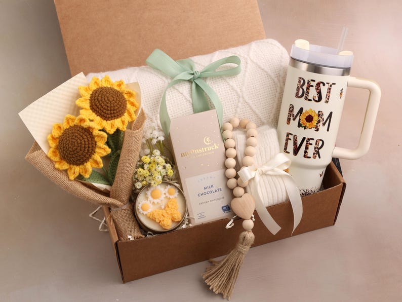 May include: A gift box filled with a white blanket, a tumbler with "BEST MOM EVER" text, a bouquet of crochet sunflowers, a wooden bead garland, a candle, and a chocolate bar. The box is tied with a green ribbon.