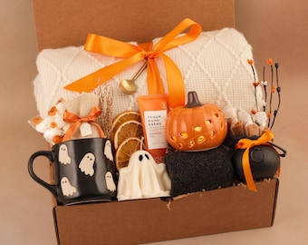 May include: A gift box containing a cream-coloured knit blanket with an orange ribbon, a black mug with white ghost patterns, a ghost-shaped candle, dried orange slices, a tube of vegan hand creme, a ceramic pumpkin, and a small black cauldron.