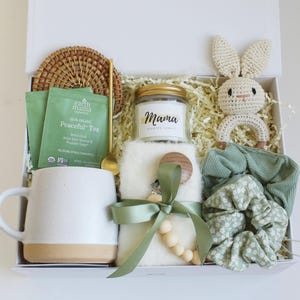Puede incluir: Una caja de regalo llena de varios art&iacute;culos. Incluye una taza blanca con base beige, una vela con la etiqueta "Mama", un conejito de ganchillo, bolsitas de t&eacute;, una cuchara, un pa&ntilde;o para eructar, un chupete, un posavasos y coleteros.