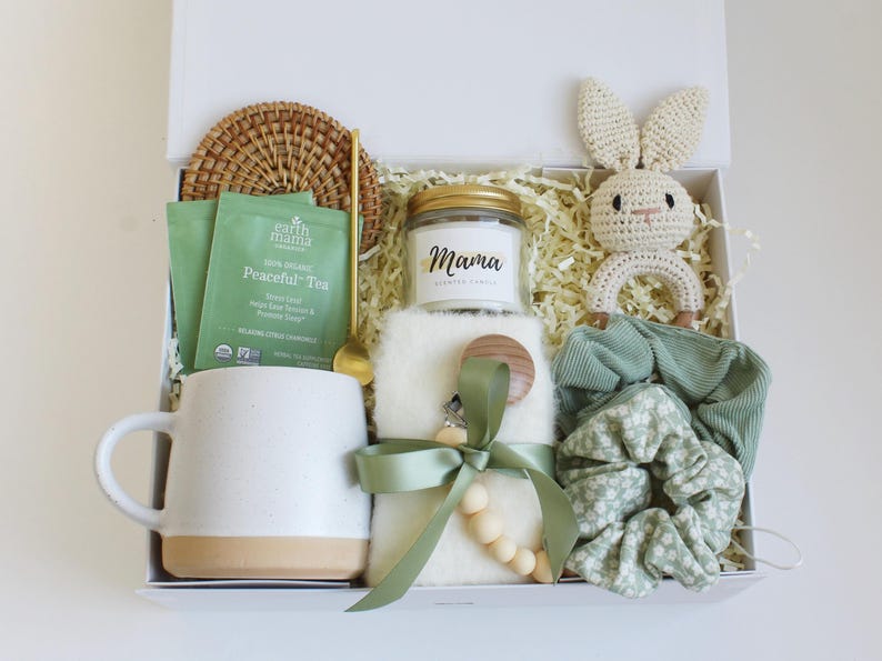 Puede incluir: Una caja de regalo llena de varios art&iacute;culos. Incluye una taza blanca con base beige, una vela con la etiqueta "Mama", un conejito de ganchillo, bolsitas de t&eacute;, una cuchara, un pa&ntilde;o para eructar, un chupete, un posavasos y coleteros.