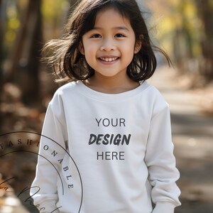 Può includere: Una bambina che indossa una maglietta bianca a maniche lunghe con la scritta "YOUR DESIGN HERE" stampata sul davanti. È in piedi in un ambiente boschivo con foglie autunnali sullo sfondo.