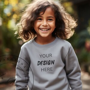 Può includere: Un bambino sorridente indossa una felpa grigia con la scritta "YOUR DESIGN HERE" in nero. La felpa è di colore uniforme e il bambino ha lunghi capelli castani ondulati. Lo sfondo è sfocato, con fogliame verde e giallo.