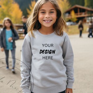 Può includere: Una felpa grigio chiaro con la scritta "YOUR DESIGN HERE" in nero. La felpa è a girocollo. Una giovane persona indossa la felpa e sorride. Lo sfondo è sfocato, con alberi e un edificio visibili.