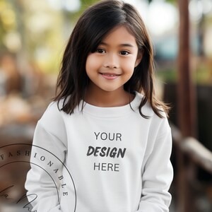 Può includere: Una persona giovane indossa una felpa bianca con la scritta "YOUR DESIGN HERE" stampata sul davanti. La felpa ha maniche lunghe e un collo rotondo.