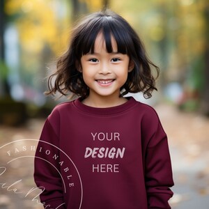 May include: A young girl wearing a maroon sweatshirt with the text "YOUR DESIGN HERE" printed on the front. She is smiling and looking at the camera.