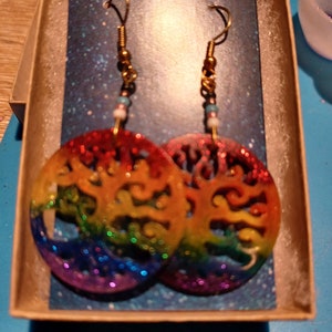 May include: A pair of rainbow glitter tree of life earrings with gold hooks and white and blue beads.