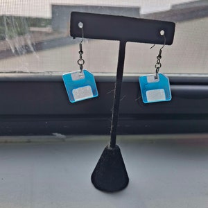 May include: A pair of silver earrings with blue and white floppy disk charms. The earrings are hanging from a black earring stand.