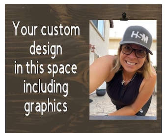 May include: A dark brown wooden picture frame with a photo held by a black clip. The frame has the text "Your custom design in this space including graphics" in white. The photo shows a person wearing a gray hat with the letters "HSM".