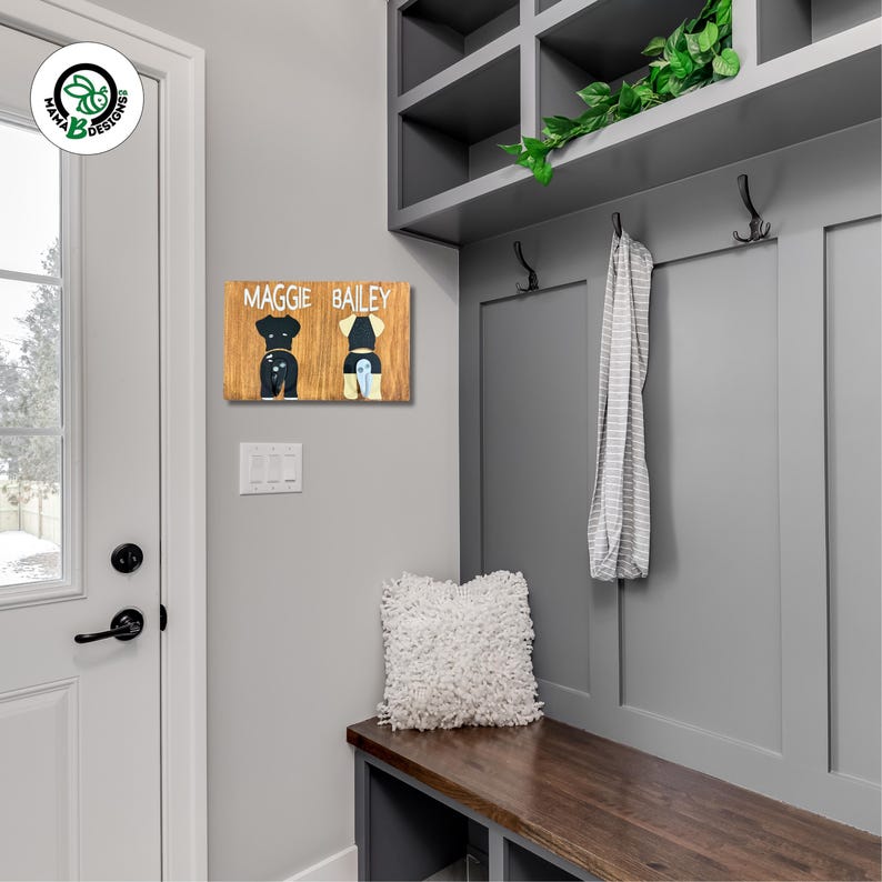 Styled photo of a double dog leash holder hanging on a home wall near a light switch. Features two painted black and white dogs with matching metal tail hooks—one black and one white. Handmade wood sign perfect for pet parents looking for personalized, functional wall decor and leash storage. Ideal gift for dog lovers and dog owners.