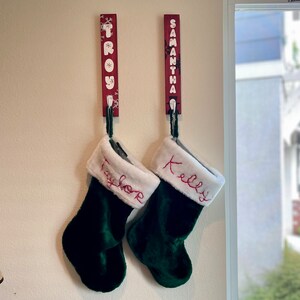 May include: Two Christmas stockings hang from the wall. Each dark green stocking has a white cuff with a name embroidered in red. Above, two red wooden nameplates read "Troy" and "Samantha" in white lettering, with snowflake accents.
