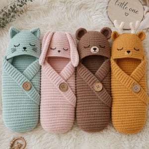 May include: Four crochet baby swaddles featuring animal hoods: a mint green cat, a pink rabbit, a brown bear, and a mustard yellow deer with antlers. Each swaddle has a single wooden button closure and embroidered closed eyes and noses.