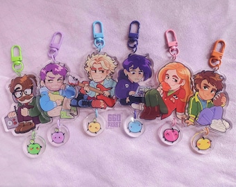 May include: Set of seven colorful keychains featuring cartoon characters with different expressions. Each keychain has a small, clear charm with a cartoon apple design. The keychains are attached to metal rings.