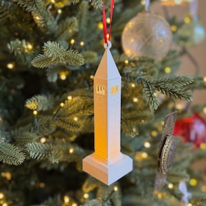 May include: White ceramic ornament shaped like a tower with a red ribbon hanging from the top. The ornament is lit from inside, creating a warm glow.