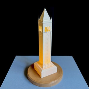 May include: A white paper model of a clock tower with a light shining through the windows. The tower is sitting on a wooden base.