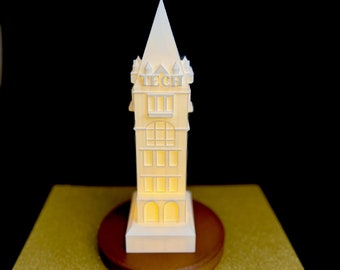 Puede incluir: Un modelo 3D impreso en blanco de un edificio con una torre de reloj. El edificio tiene un letrero que dice "TECH" en la parte delantera. El modelo está iluminado desde adentro y está sentado sobre una base de madera.