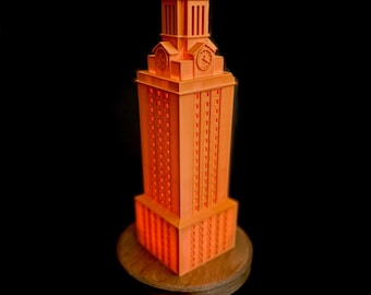 May include: A miniature replica of the University of Texas at Austin's iconic tower, the UT Tower, made of orange plastic. The tower is on a wooden base.