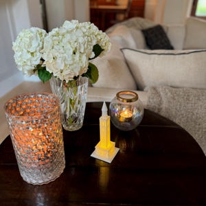 May include: A white ceramic tower with a yellow light inside, a clear glass candle holder with a gold rim and a lit candle inside, and a clear glass vase with white hydrangeas on a dark wood table.