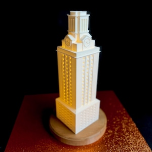 May include: A white 3D printed model of the University of Texas at Austin's iconic tower, the UT Tower, with a clock face and illuminated windows. The model is on a wooden base.